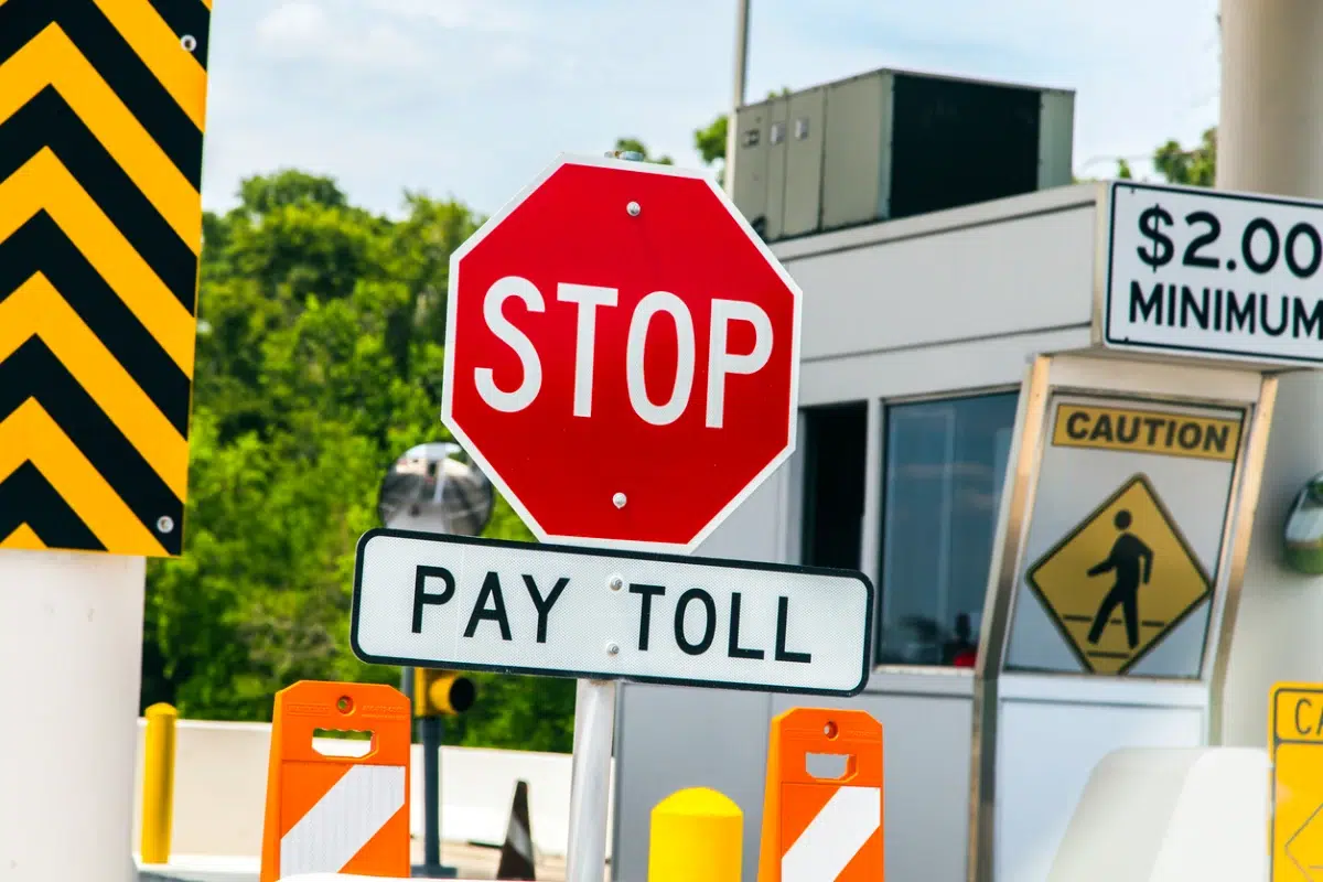 A toll booth with a red stop sign reading "Pay Toll," a $2.00 minimum sign, caution pedestrian sign, and orange barriers.