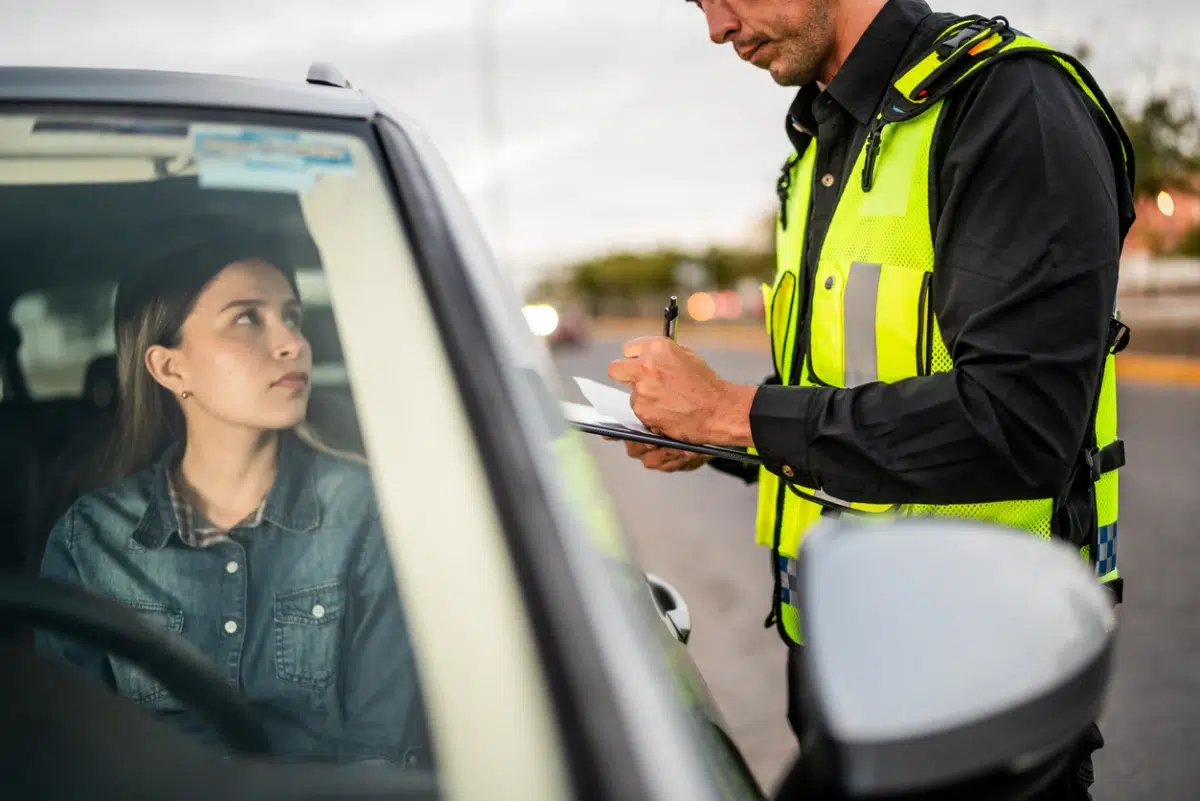 A police officer in a high-visibility vest writes a ticket while a young woman sits in her car during a traffic stop.