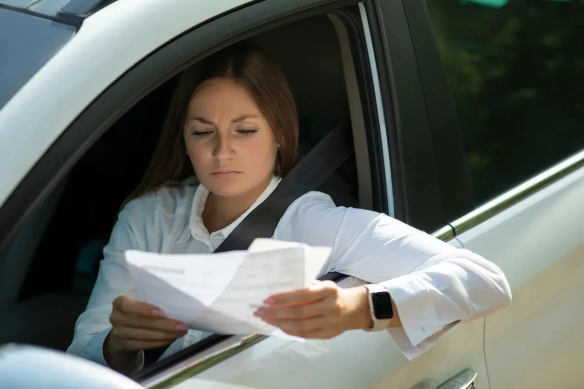 Woman sitting in a car reading documents with a serious expression, wearing a white shirt and smartwatch.