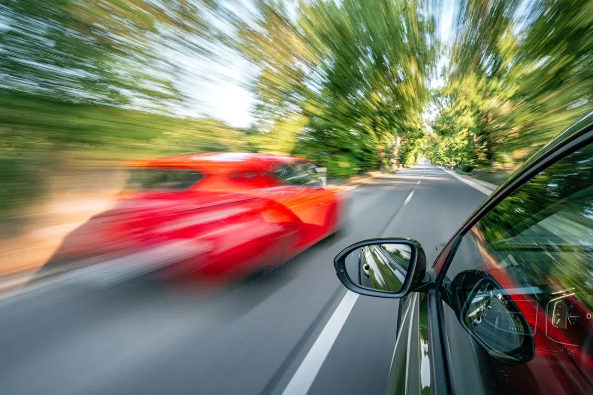 Two cars speeding on a tree-lined road, motion blur effect showing a red car passing a dark vehicle.