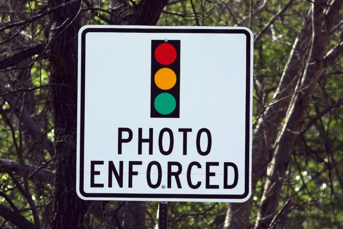 White traffic sign with a stoplight graphic reading "Photo Enforced," mounted outdoors with trees in the background.