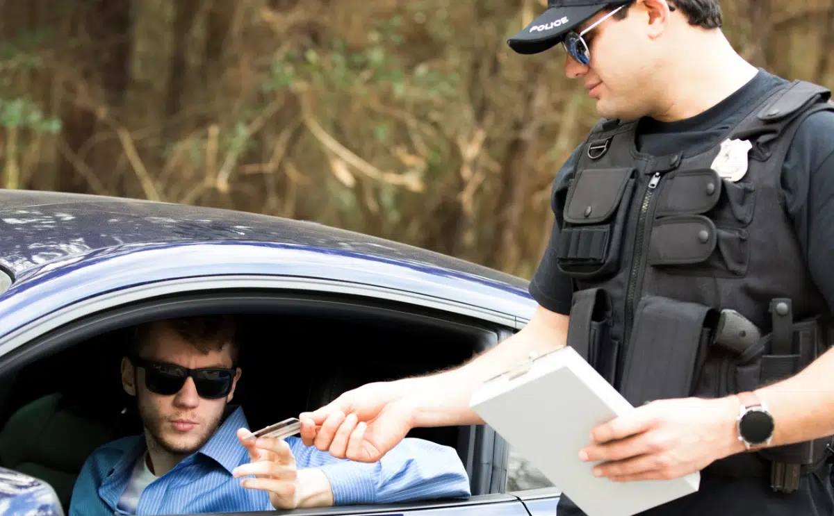 A police officer holding a clipboard receives documents from a driver during a traffic stop.