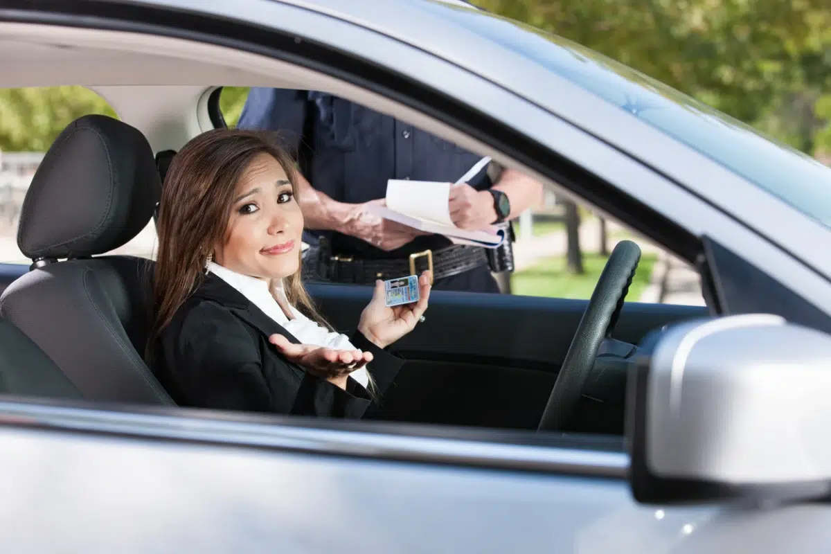 Woman in car holding her driver's license with a nervous expression while a police officer writes a ticket outside.