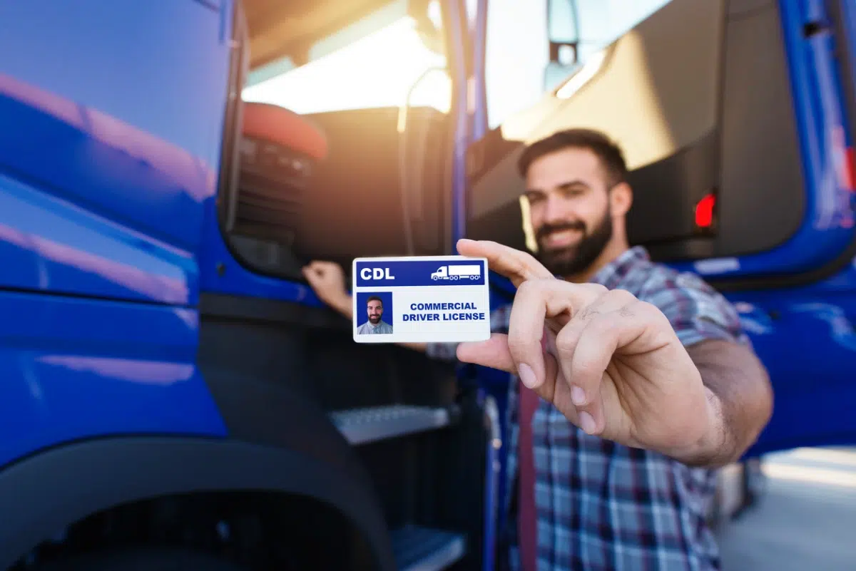 Smiling bearded man holding a CDL Commercial Driver License card in front of a blue semi-truck.
