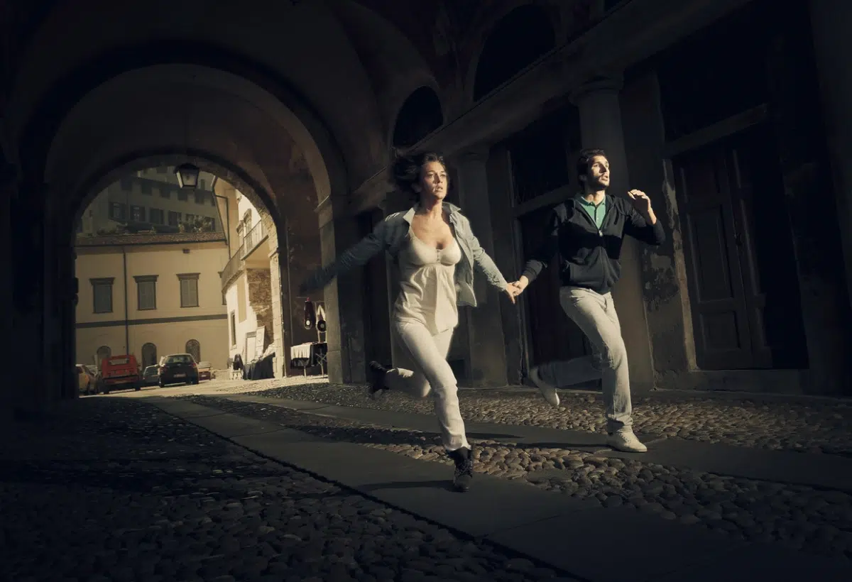 A couple runs hand-in-hand through a dark cobblestone archway in a European city street.
