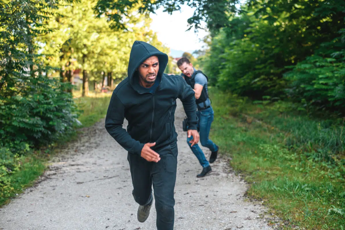 A man in a black hoodie sprinting down a tree-lined path while another man in a vest chases him from behind.